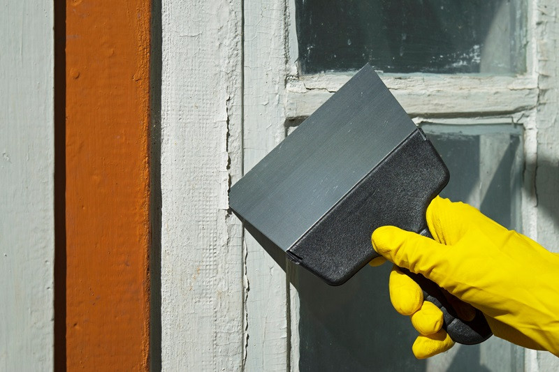 Renovation of an old house. A worker’s hand in a yellow glove cleans paint from a window with a spatula. Removing the coating with a construction tool. Household renovation. Selective focus